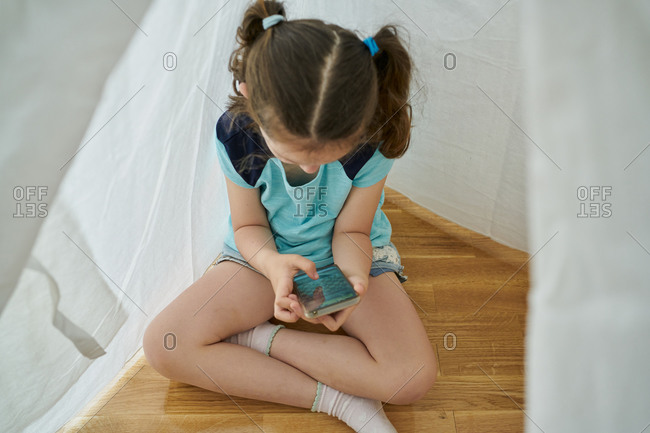 Little girl looking at her smartphone inside a white teepee tent inside her house.
