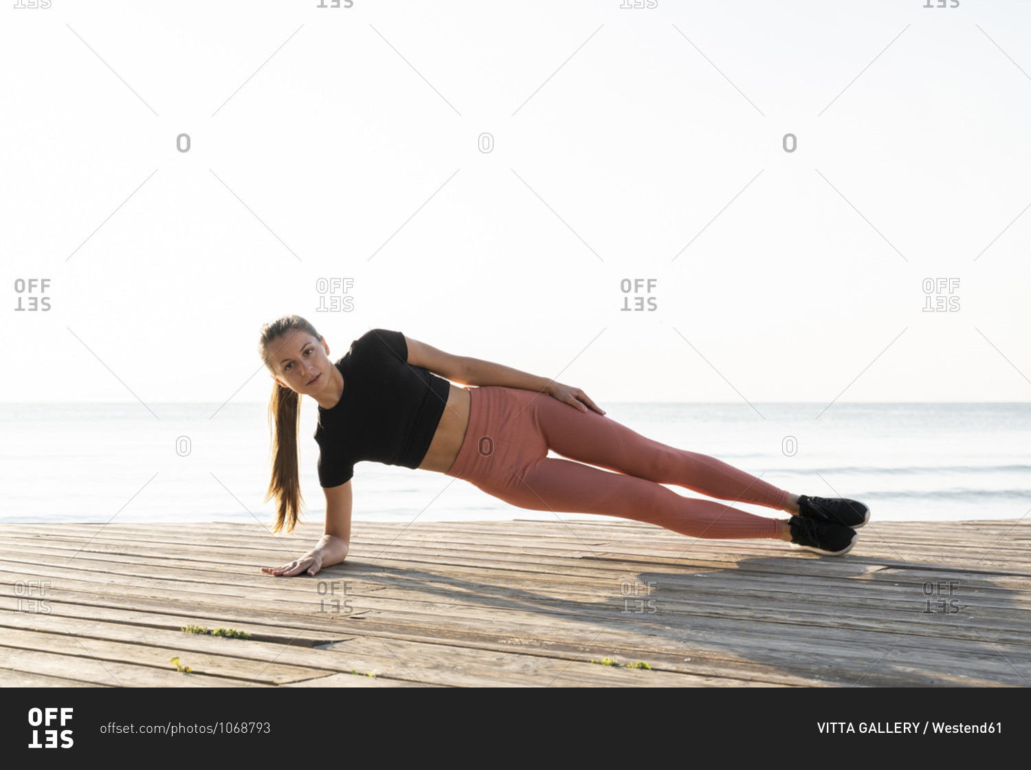 Young sportswoman doing side plank pose on promenade against clear sky
