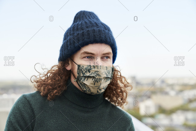 Thoughtful with curly long face mask and beanie on rooftop stock photo - OFFSET