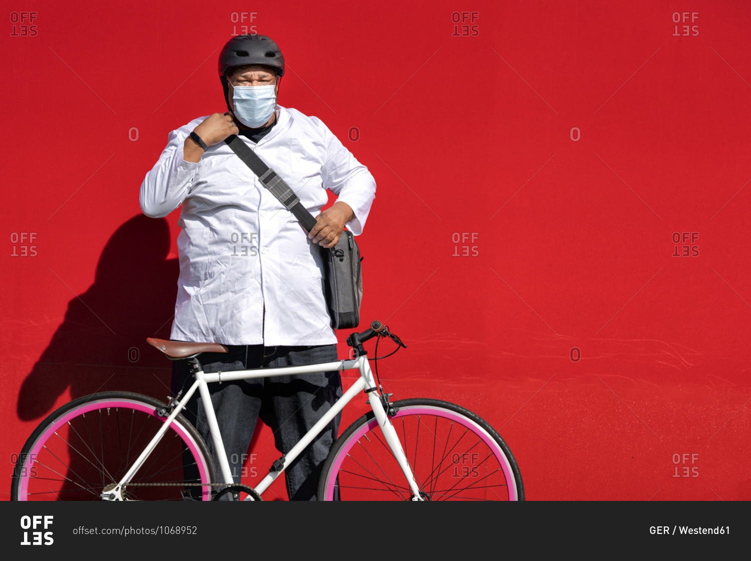 Mature man wearing protective face mask standing with bicycle against red wall on sunny day