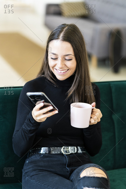 Smiling young female influencer holding smart phone and coffee cup while sitting on sofa at loft apartment