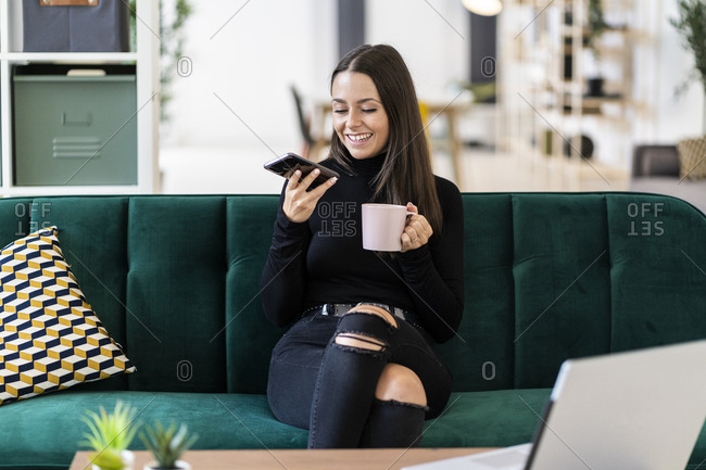 Smiling young female blogger holding smart phone and coffee cup while sitting on sofa at loft apartment