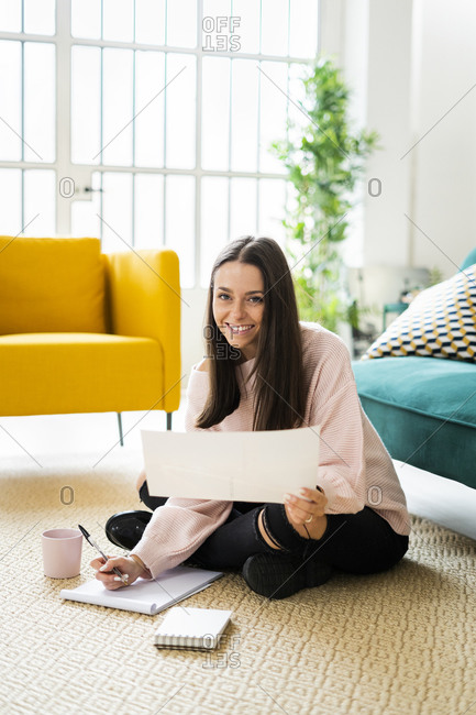Happy beautiful young woman sitting with note pads and coffee cup on carpet against sofa at home