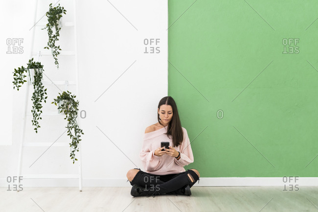 Beautiful young woman text messaging through mobile phone while sitting on floor by ladder against wall