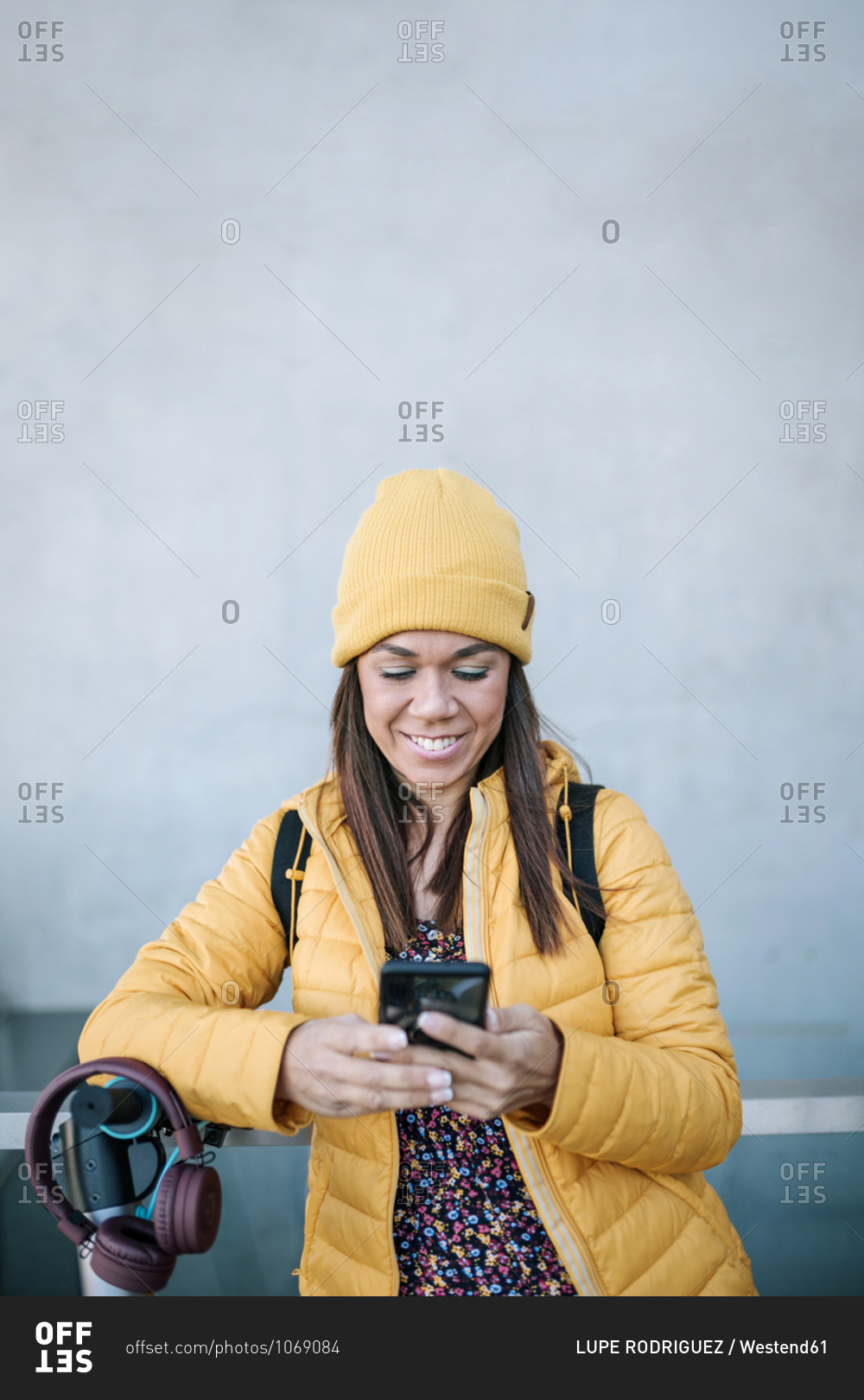 Smiling woman using mobile phone while leaning on electric push scooter against wall