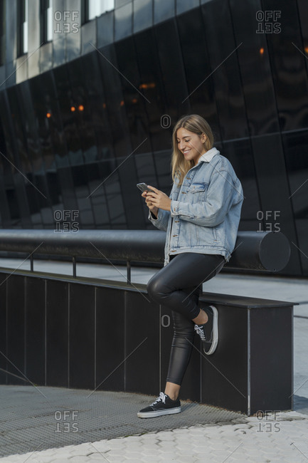 Smiling young woman using smart phone while leaning on railing outside modern building
