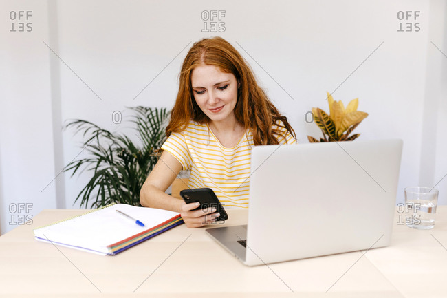 Redhead woman text messaging on smart phone sitting by laptop at home