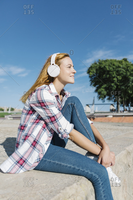Woman listening music through headphone looking away while sitting on footpath during sunny day