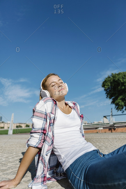 Woman with eyes closed listening music through headphone sitting footpath during sunny day