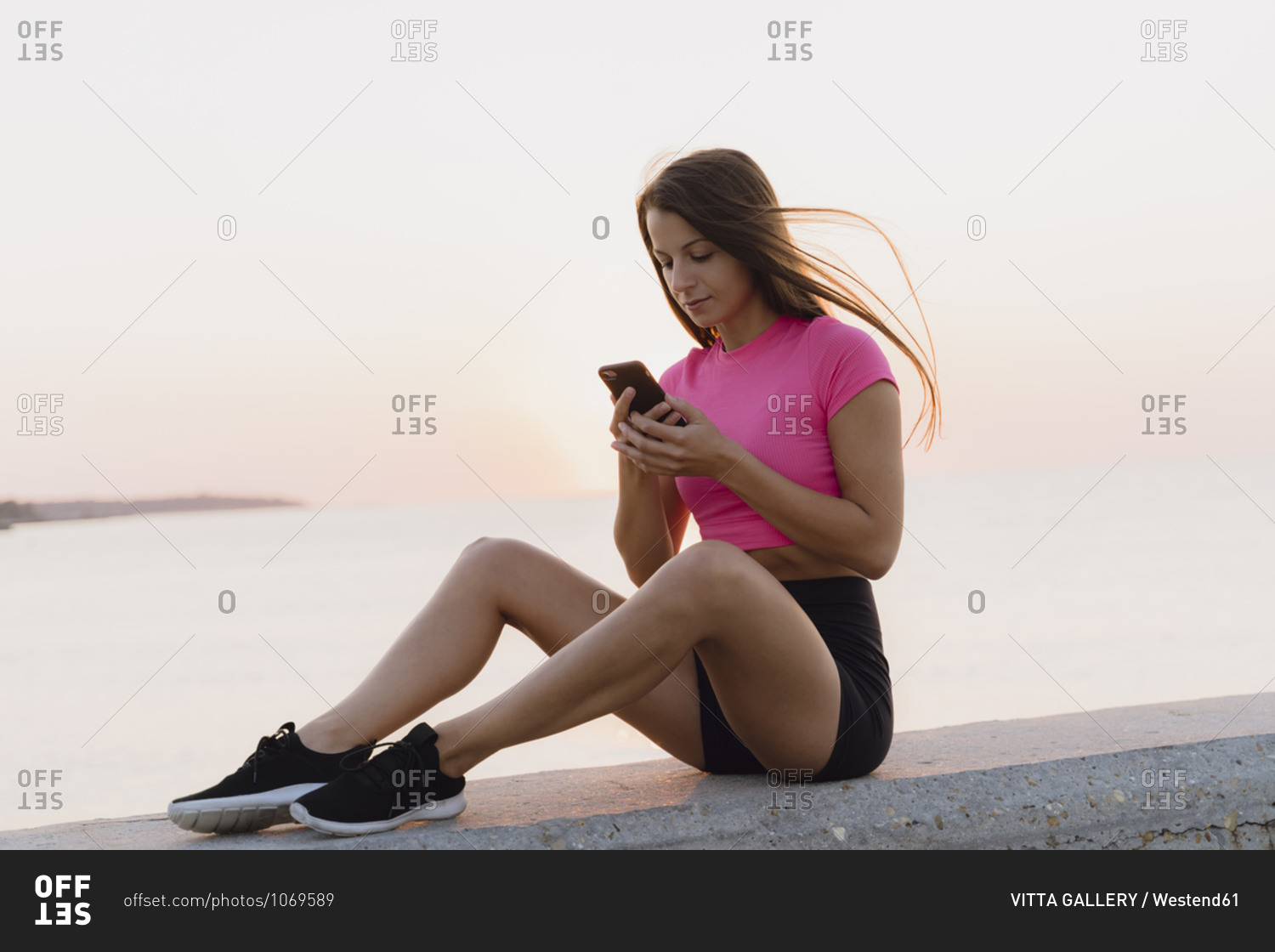 Athlete using mobile phone while sitting on concrete wall against sea