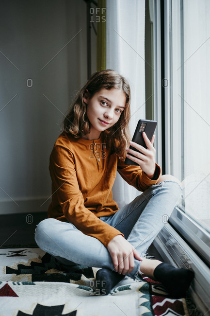 Girl with mobile phone sitting cross legged by window at home
