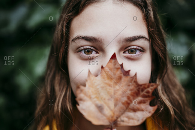 Girl in raincoat hiding face with dry ivy leaf while standing against leaf wall