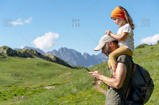 Father using smart phone while daughter sitting on his shoulder during sunny day