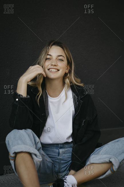 Smiling young woman sitting on footpath against wall