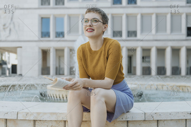 Mid adult woman looking away with book sitting against fountain