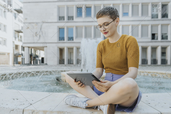 Woman with cross legged using digital tablet sitting by fountain on sunny day