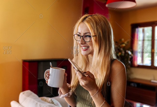 Smiling girl enjoying coffee while holding mobile phone at home