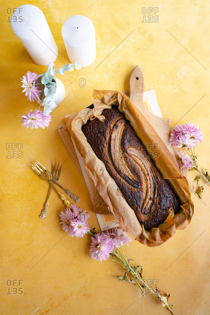 Whole loaf of banana bread with no added sugar in baking pan with parchment paper on yellow surface with candles and flowers