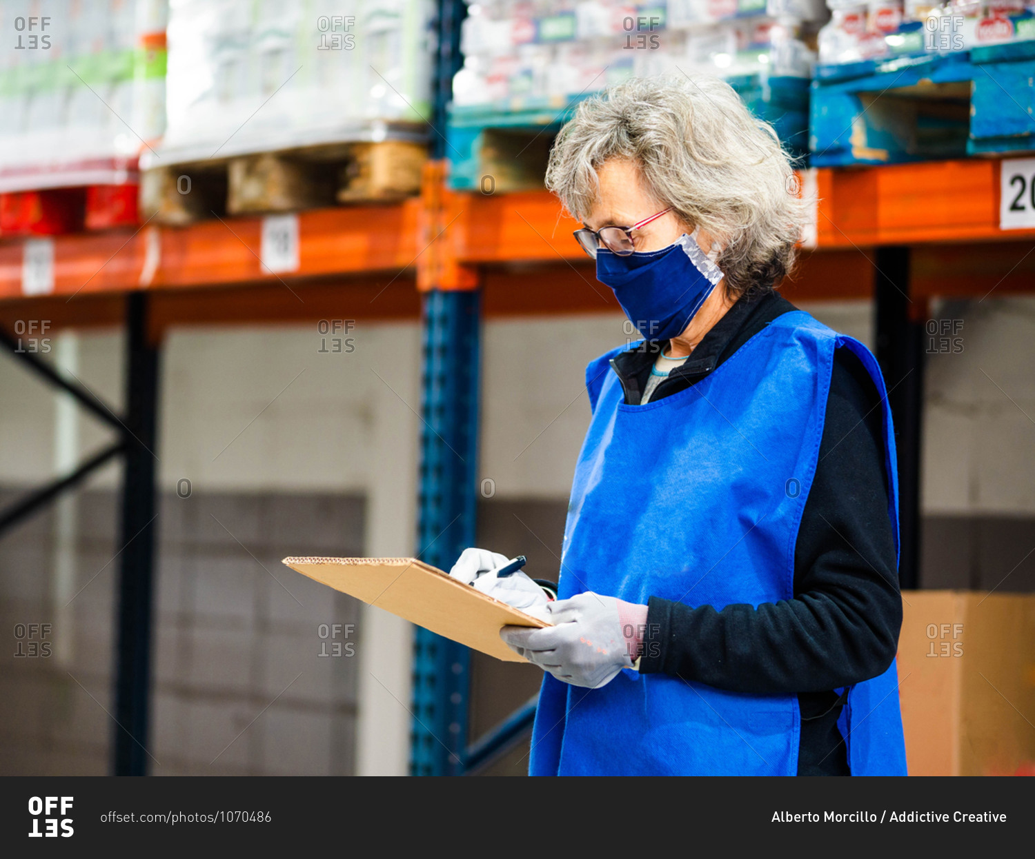 Side view focused female worker in blue vest and face mask taking notes in clipboard while standing near stacks in warehouse