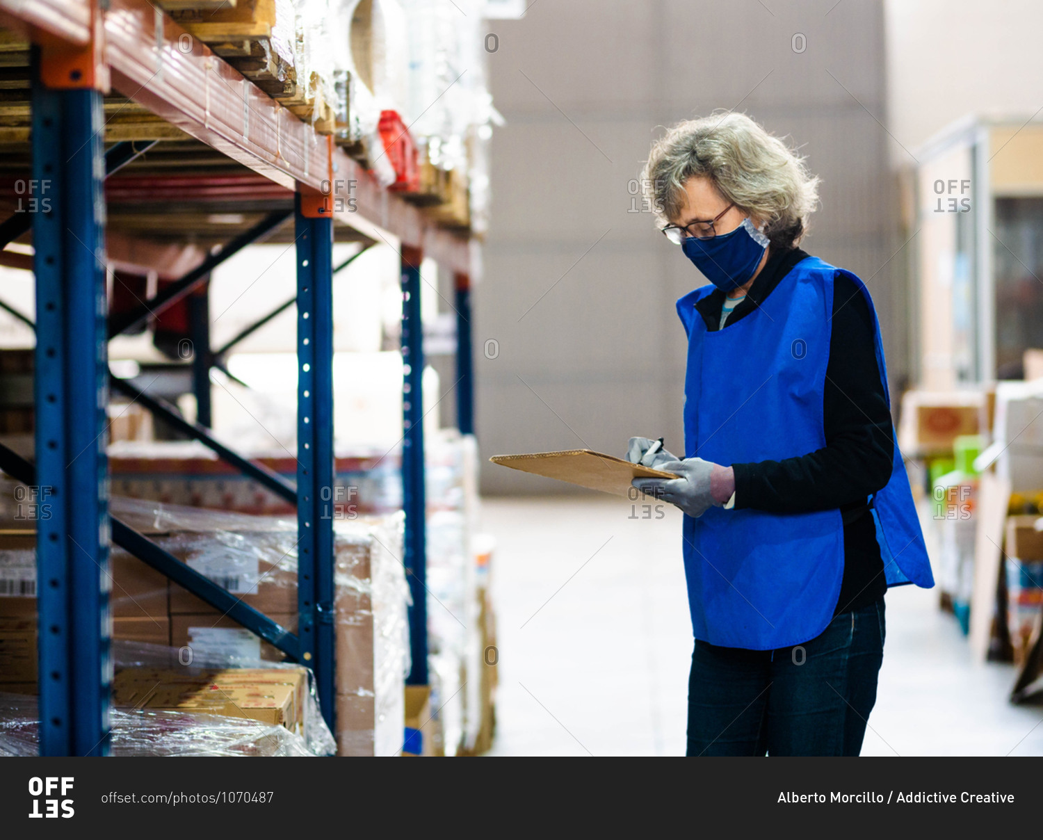 Side view focused female worker in blue vest and face mask taking notes in clipboard while standing near stacks in warehouse
