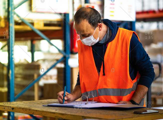 Side view adult worker wearing uniform and protective face mask writing on clipboard while working in spacious storehouse