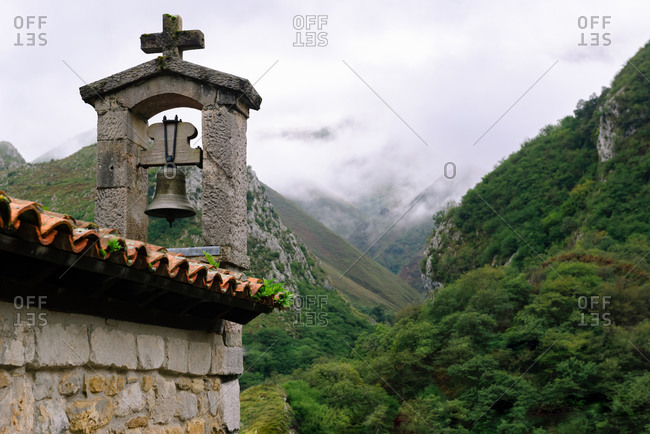 Greenery ridge in mist near aged masonry temple with bell and cross under cloudy sky in Spain