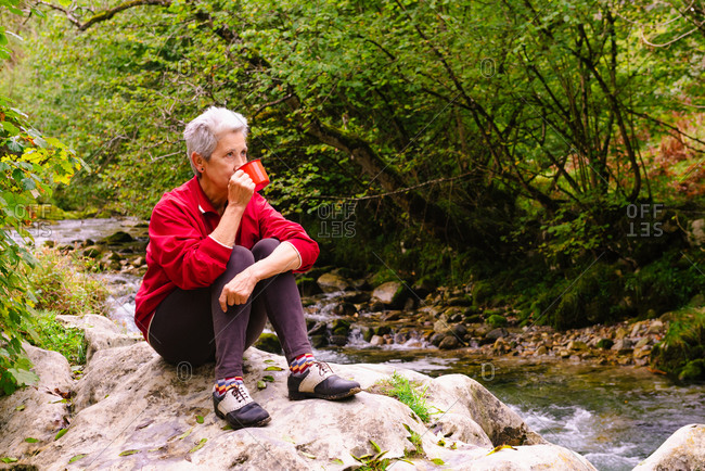 Contemplative elderly female tourist drinking water near Casano river while resting on stone and looking away