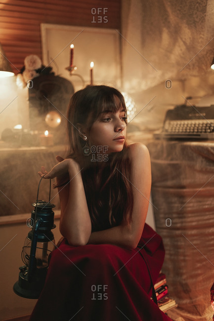 Peaceful female in red dress sitting in cozy room with old fashioned kerosene lantern while enjoying evening and looking away
