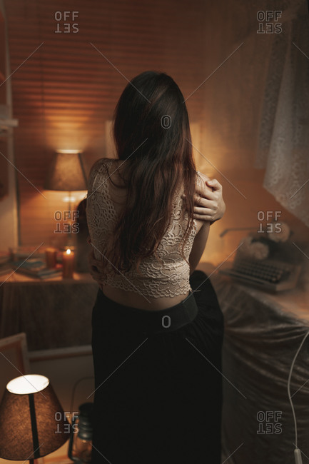 Back view of anonymous female in lace bra embracing herself while sitting in cozy room lit by dim orange light