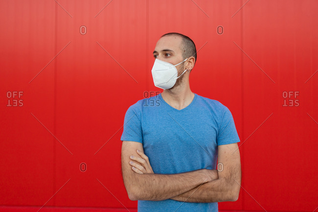Young man wearing respiratory mask with arms crossed while looking away near colorful red wall during coronavirus pandemic