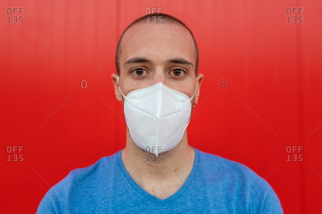 Young man wearing respiratory mask while looking at camera near colorful red wall during coronavirus pandemic