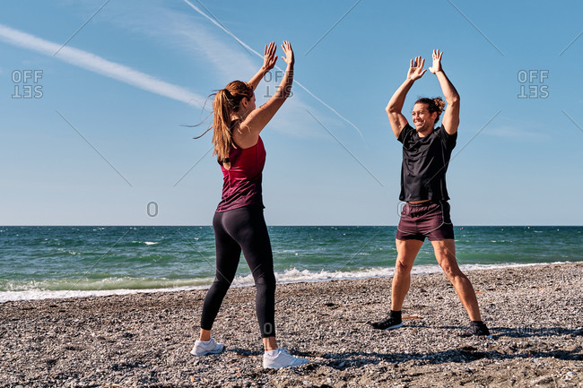 Sporty energetic female doing exercises with raised arms under supervision of personal instructor while standing on beach in summer and looking at each other