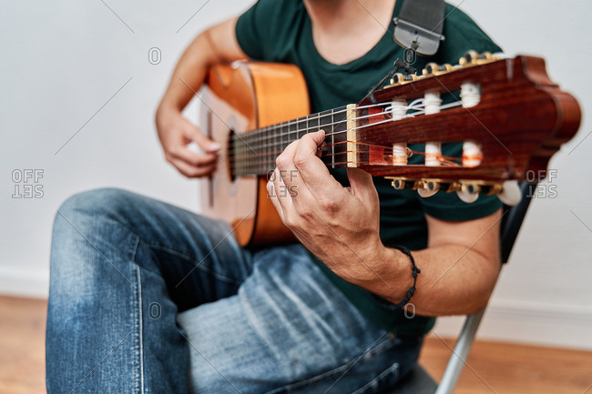 Cropped unrecognizable focused male guitarist in jeans playing acoustic guitar while sitting on chair at home