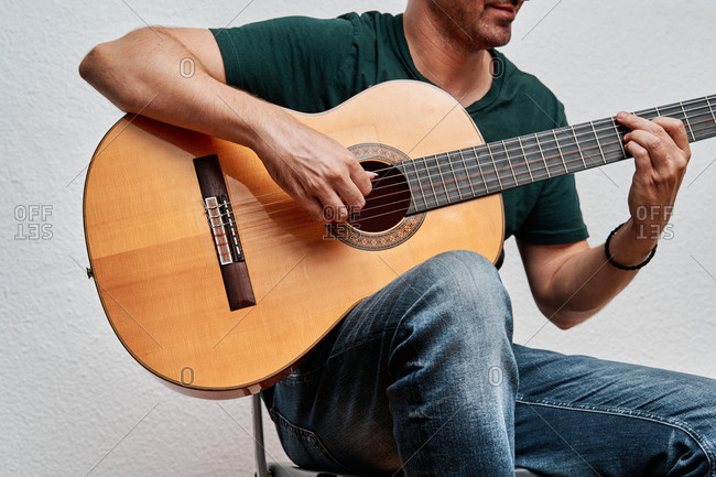 Cropped unrecognizable focused male guitarist in jeans playing acoustic guitar while sitting on chair at home on white background