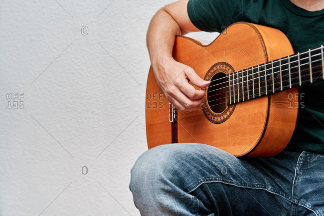 Cropped unrecognizable focused male guitarist in jeans playing acoustic guitar while sitting on chair at home on white background