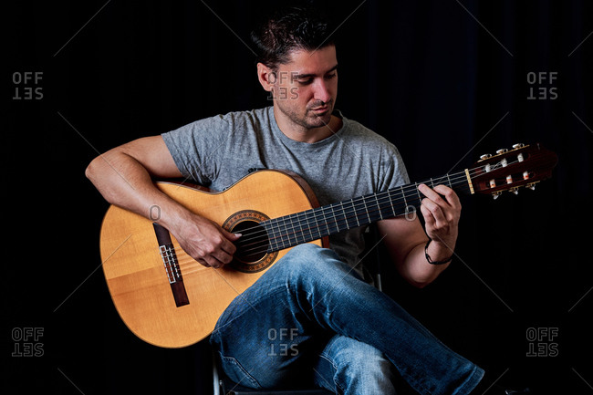 Focused male guitarist in jeans playing acoustic guitar while sitting on chair at home