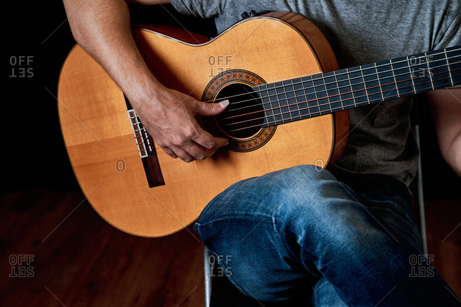Cropped unrecognizable focused male guitarist in jeans playing acoustic guitar while sitting on chair at home