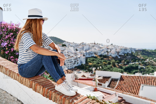 Side view of female tourist in sunhat and casual wear sitting on stone border and enjoying cityscape of Frigiliana while embracing knees and looking away