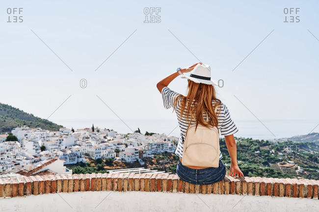 Back view of delighted female tourist sitting on stone border enjoying freedom on background of cityscape of Frigiliana during vacation in summer