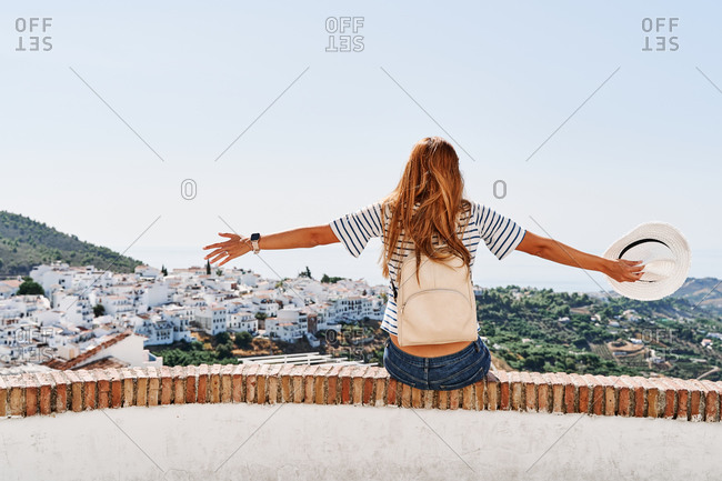 Back view of delighted female tourist sitting on stone border with outstretched arms and enjoying freedom on background of cityscape of Frigiliana during vacation in summer