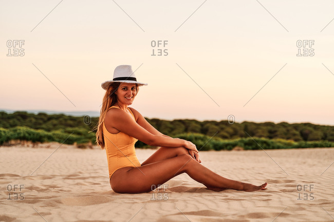 Side view of anonymous delighted female wearing swimwear sitting and looking at camera while enjoying freedom on sandy shore