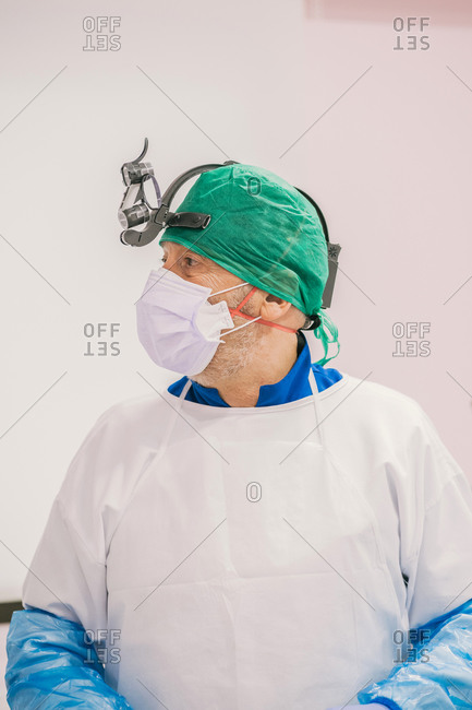 Unrecognizable male stomatologist in protective mask and binoculars looking away in clinic near wall
