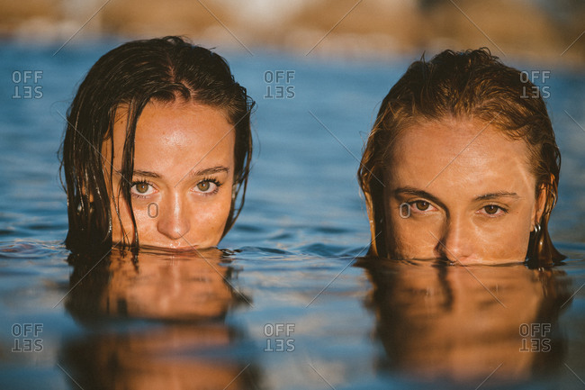 Charming female friends covering mouths in sea water while looking at camera at sunset during vacation in summer