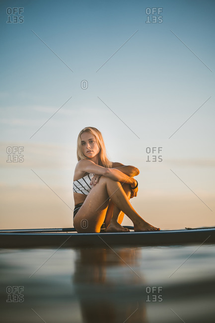 Side view low angle of slim female in swimsuit sitting on SUP board in sea and embracing knees while looking at camera and enjoying sunset