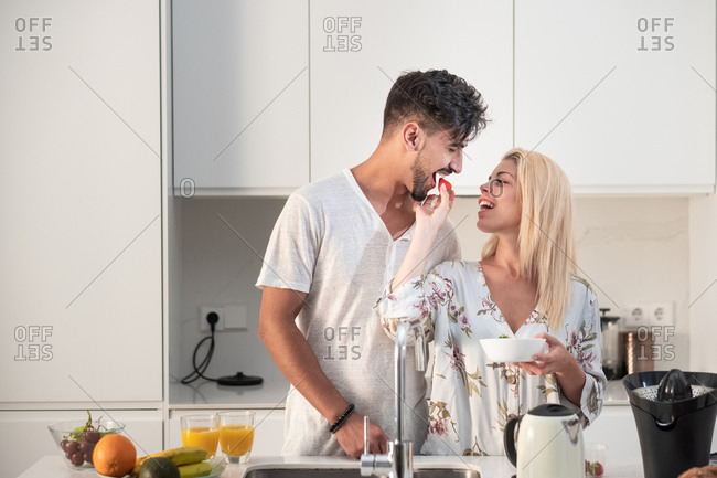 Tender couple eating strawberries and drinking fresh orange juice while hugging in kitchen in morning during breakfast