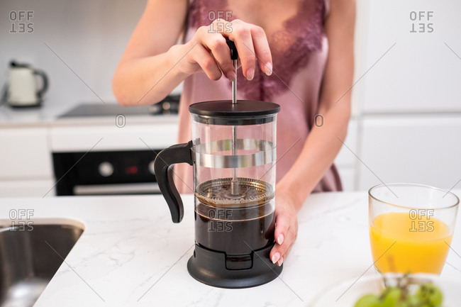 Unrecognizable female in silk pajama standing at counter in kitchen and preparing aromatic coffee in French press