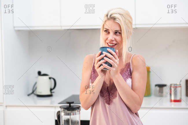 Calm female in silk pajama standing at table in modern kitchen and enjoying fresh morning coffee at breakfast at home