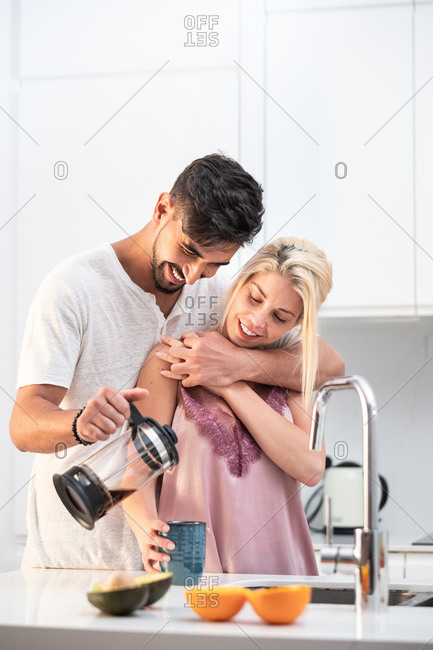 Loving couple in pajamas hugging and pouring fresh coffee in cup together while standing at table in kitchen during breakfast in morning