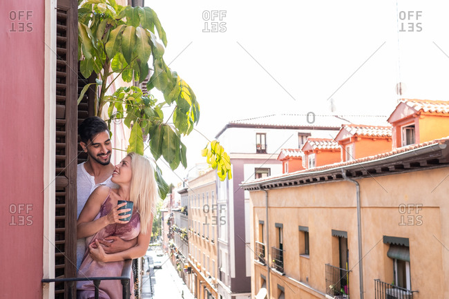 Smiling man cuddling gentle woman with cup of coffee while standing together on balcony in morning and looking at each other