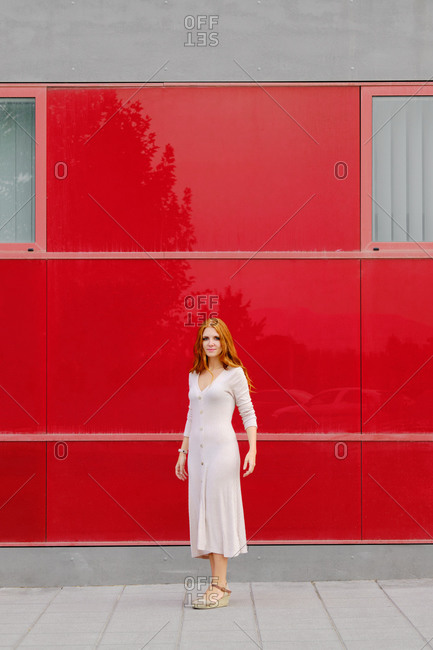 Full body of young redhead female in elegant beige dress standing on pavement against bright red wall of modern urban building